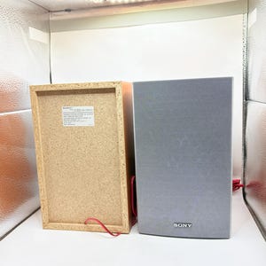 May include: Two Sony speakers, one showing the light brown particle board back with a white identification label and red wire, and the other showing the grey fabric front grille with a silver Sony logo at the bottom and a red wire extending.