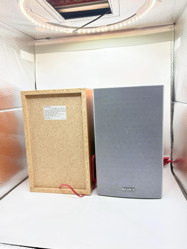 May include: Two Sony speakers, one showing the light brown particle board back with a white identification label and red wire, and the other showing the grey fabric front grille with a silver Sony logo at the bottom and a red wire extending.