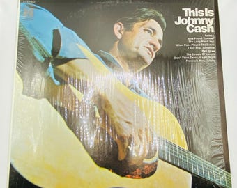 May include: Vintage vinyl record album cover featuring Johnny Cash playing a guitar. The album is titled "This Is Johnny Cash" and includes a list of songs. The cover art shows Cash in a blue shirt, with a yellow guitar.