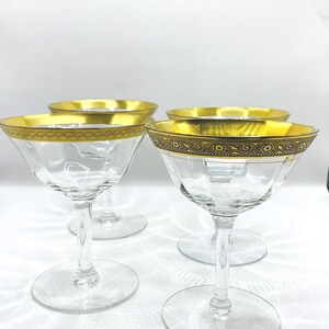 Könnte beinhalten: Vier klare Champagnergläser mit goldenen Rändern und dekorativen Details. Die Gläser haben eine klassische Coupe-Form mit einer breiten Schale und einem schlanken Stiel. Der goldene Rand verleiht dem Glaswaren einen Hauch von Eleganz.