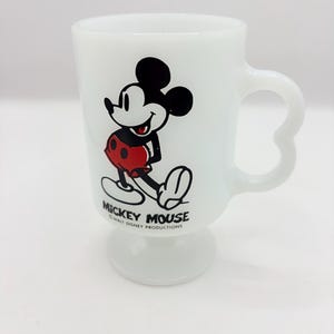 May include: A white milk glass pedestal mug featuring a graphic of Mickey Mouse in red shorts. The text MICKEY MOUSE and WALT DISNEY PRODUCTIONS is printed in black below the character. The mug has a curved handle and a circular base.