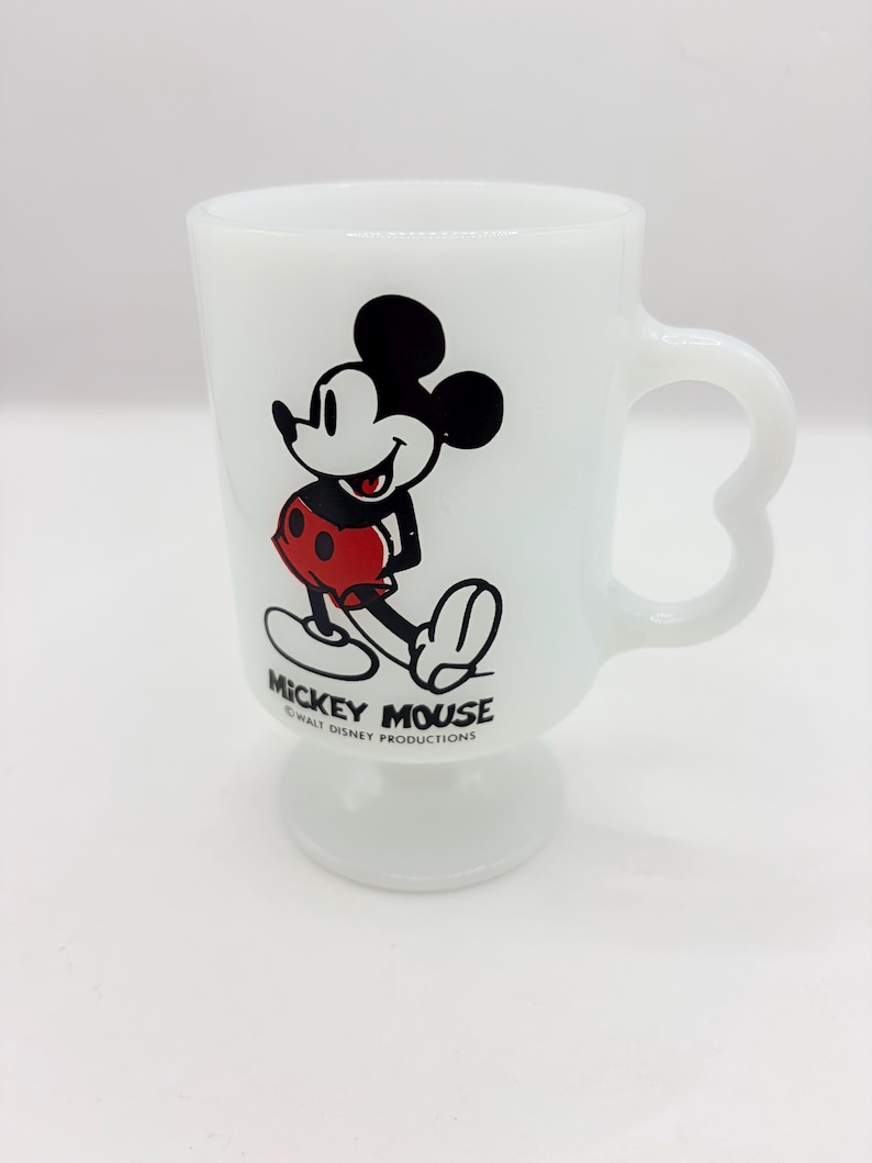 May include: A white milk glass pedestal mug featuring a graphic of Mickey Mouse in red shorts. The text MICKEY MOUSE and WALT DISNEY PRODUCTIONS is printed in black below the character. The mug has a curved handle and a circular base.