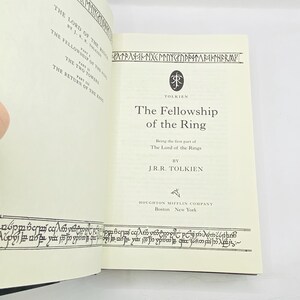 May include: Open book with the title "The Fellowship of the Ring" by J.R.R. Tolkien. The book's pages are cream-colored with black text and decorative borders. The cover is not visible. The book is part of "The Lord of the Rings" series.
