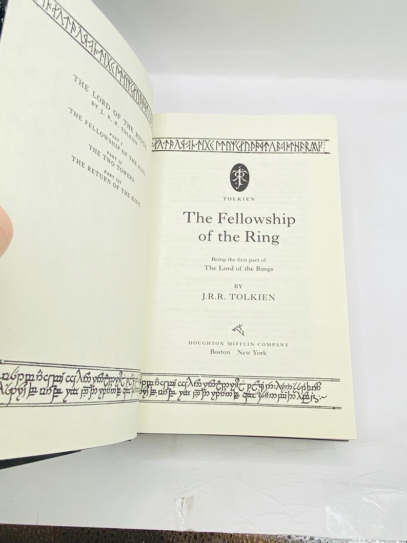 May include: Open book with the title "The Fellowship of the Ring" by J.R.R. Tolkien. The book's pages are cream-colored with black text and decorative borders. The cover is not visible. The book is part of "The Lord of the Rings" series.