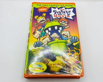 Pode incluir: Uma fita VHS de "The Rugrats Movie" com uma capa colorida com personagens de desenhos animados. A fita está em uma embalagem plástica transparente, com texto que diz "THE FULL-LENGTH HIT MOVIE" e "...a delight!"