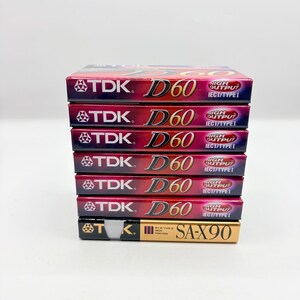 May include: A vertical stack of seven audio cassette tapes. The top six tapes are TDK D60 models with red and black packaging, featuring the text TDK D60 High Output IEC I/Type I. The bottom tape is a gold and black TDK SA-X90 High Position cassette.