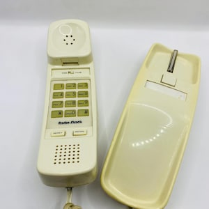 May include: Off-white vintage Radio Shack telephone with a coiled cord. The phone has a keypad with numbers and function buttons, including "RESET" and "REDIAL". The base has a cradle for the handset.