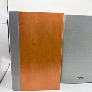 May include: A pair of rectangular speakers featuring a smooth, light brown wood grain finish on the front panel. Each speaker has a vertical strip of grey mesh fabric along the side. The right speaker displays the Sony brand name in silver text.