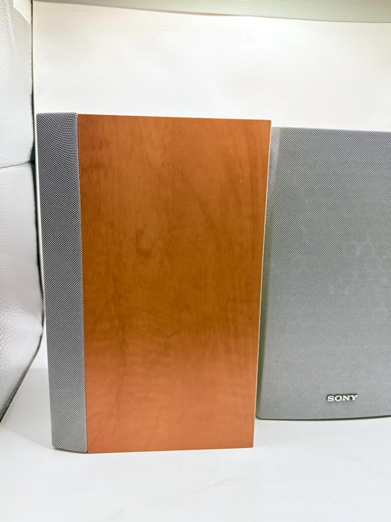 May include: A pair of rectangular speakers featuring a smooth, light brown wood grain finish on the front panel. Each speaker has a vertical strip of grey mesh fabric along the side. The right speaker displays the Sony brand name in silver text.
