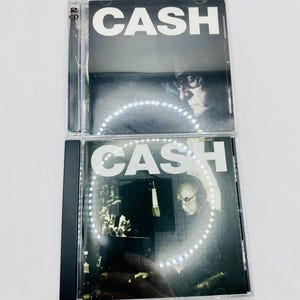 May include: Two CD cases with the word "CASH" in large white letters. The CD covers feature black and white images of a man with glasses. The top CD case has "2 CD" printed on the spine.