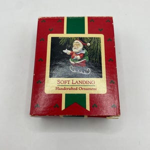 Peut inclure: Boîte d'ornement de Noël rouge et verte avec une image du Père Noël sur un traîneau. La boîte porte l'inscription "SOFT LANDING Handcrafted Ornament". La boîte est rectangulaire et comporte une bande verte sur le dessus.