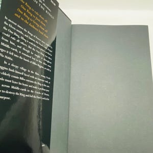 May include: Open book with a black cover featuring the title "The Lord of the Rings" and text from the book. The pages are gray. The book is open to reveal the inside pages.