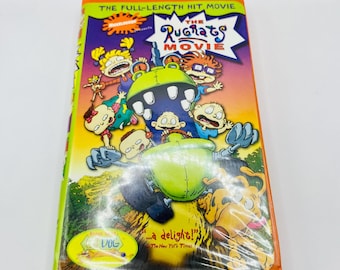 Pode incluir: Uma fita VHS de "The Rugrats Movie" com uma capa colorida com personagens de desenhos animados. O título está em uma fonte estilizada e a fita é selada em plástico. A capa também inclui o texto "...a delight!" e "The New York Times."