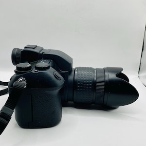 Puede incluir: Cámara réflex digital negra con objetivo zoom y parasol. La cámara tiene una correa adjunta y varios botones y diales. La cámara está sobre una superficie blanca.