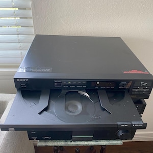 Puede incluir: Un reproductor de CD Sony negro con una bandeja de disco abierta. El reproductor tiene una pantalla digital y botones de control. La unidad está sobre una mesa pequeña con base de madera oscura. Componente de audio vintage.