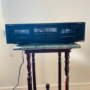 Könnte beinhalten: Schwarzes Onkyo-Kassetten-Deck auf einem kleinen Tisch aus dunklem Holz mit einer grünen Marmorplatte. Das Deck hat mehrere Knöpfe und Anzeigen, mit dem Onkyo-Logo. Der Tisch hat verzierte Beine.