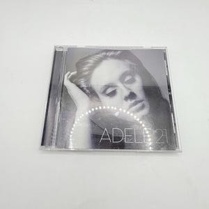May include: A square compact disc case featuring a black and white portrait of a person with their hand near their head. The text ADELE 21 is printed in white capital letters across the lower portion of the cover art, which is encased in clear plastic.
