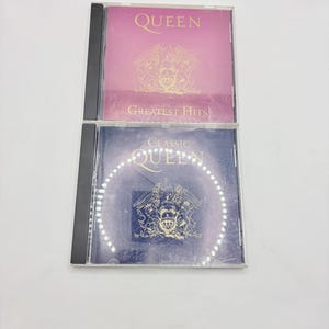 May include: Two Queen music CDs. The top CD case is pink with the word "QUEEN" and "GREATEST HITS" in white text. The bottom CD case is navy blue with the words "CLASSIC QUEEN". Both cases feature a gold emblem.