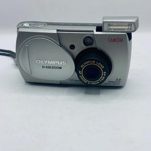 Könnte beinhalten: Silberne Olympus D-520 Zoom Digitalkamera mit ausklappbarem Blitz. Die Kamera hat ein schwarzes Objektiv und die Texte "OLYMPUS" und "CAMEDIA" sind sichtbar. Das Objektiv ist mit "AF Zoom 5-15mm 1:2.8-4.9" beschriftet.