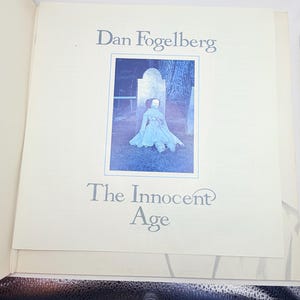 May include: A vinyl record gatefold cover featuring the text Dan Fogelberg and The Innocent Age. The center image shows a doll in a light-colored dress sitting on the ground in front of a tombstone. The background is a cream-colored paper.