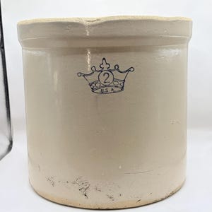 May include: A cylindrical beige stoneware crock with a thick rim and a flat base. The front features a blue stamped graphic of a crown containing the number 2 and the text U.S.A. The surface has a smooth, glazed finish with minor wear near the base.