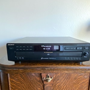 Könnte beinhalten: Ein schwarzer Sony CD-Player mit dem Text "no disc" auf dem Bildschirm. Der CD-Player steht auf einem Holzschrank. Der CD-Player hat mehrere Tasten und eine CD-Wechsler-Funktion.