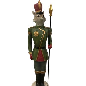 May include: A decorative fox figurine in a military uniform. The fox wears a green jacket with gold trim, a burgundy hat, and black boots. It holds a spear with a gold tip. The figurine stands on a round, wooden base.