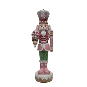 Peut inclure: Une figurine de casse-noisette de Noël rose et verte tenant une canne en sucre d'orge. Le casse-noisette porte une veste rose et rouge avec des bordures blanches et un chapeau blanc avec un motif de canne en sucre d'orge. Le casse-noisette est debout sur une base brune avec du glaçage blanc et des pépites.