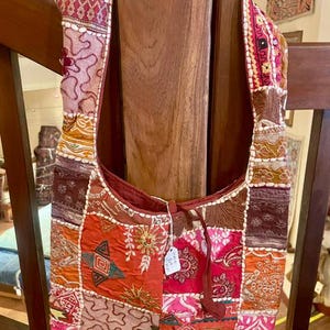 May include: A hobo-style shoulder bag crafted from a patchwork of multicolored fabric squares. The bag features intricate embroidery, floral motifs, and geometric patterns in shades of red, orange, pink, and brown, with a small white tag attached.
