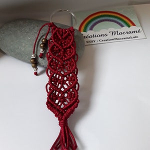 Op de afbeelding: Een rode macramé sleutelhanger met een kwast en kralen. De sleutelhanger is bevestigd aan een zilveren ring. De tekst "créations Macramé" en "ETSY - CreationMacrameLolo" is zichtbaar op een kaart achter de sleutelhanger.