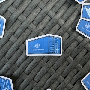 Könnte beinhalten: Ein blauer Aufkleber mit einem stilisierten Seecontainer, dem Wort "Kubernetes" und einem weißen Logo. Der Aufkleber ist rechteckig mit weißem Rand und liegt auf einem strukturierten grauen Hintergrund.