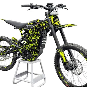 May include: Electric dirt bike in black and neon yellow, with a bold geometric design. The bike has a black seat, tyres, and handlebars, and is on a white stand. The wheels have a diameter of approximately 50 cm.