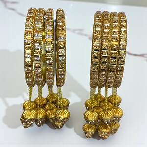 Könnte beinhalten: Zwei Sets goldfarbener Armreifen mit aufwendigen Details. Jeder Armreif ist mit kleinen, funkelnden Verzierungen versehen und verfügt über baumelnde goldene Quasten. Die Armreifen werden auf einer weißen Oberfläche präsentiert, die ihr verziertes Design hervorhebt.