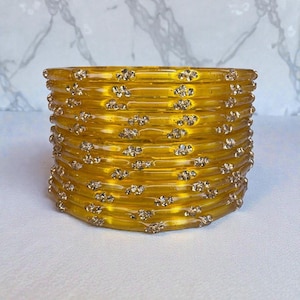 May include: A stack of yellow bangles with a faceted design. Each bangle is adorned with small, sparkling embellishments. The bangles are stacked on top of each other, creating a cylindrical shape. The background is a light, neutral color.