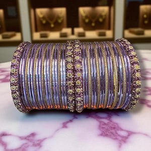 Puede incluir: Un conjunto de pulseras moradas y doradas. Las pulseras están apiladas, formando una forma cilíndrica. El diseño presenta bandas alternas de morado y dorado sólido, con extremos decorativos.
