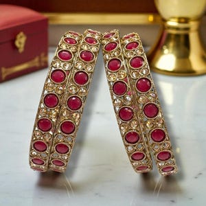 May include: Four gold bangles with red gemstones and clear accent stones. The bangles are arranged in pairs, showcasing their intricate design. The jewelry is set against a neutral background, highlighting the vibrant colors and details.