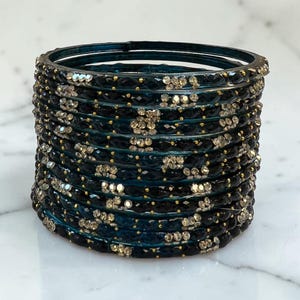 May include: A stack of twelve circular bangles featuring a dark teal and black faceted surface. Each bangle is adorned with small, clear, reflective crystals arranged in clusters and accented with tiny gold-coloured beads along the edges.