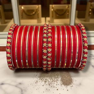May include: A collection of red bangles with gold and rhinestone embellishments. The bangles are a bright red hue, featuring gold stripes and decorative gold and rhinestone detailing on the edges and centre.