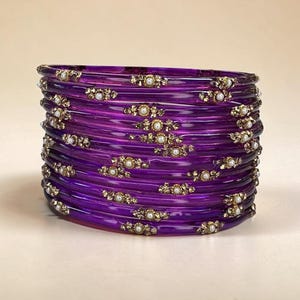 May include: A stack of translucent purple bangles featuring small, gold-toned floral embellishments. Each individual bracelet is accented with a single white pearl at the centre of the gold-toned floral design, creating a repeating pattern.
