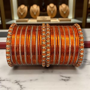 May include: A stack of orange bangles with silver accents. The bangles are adorned with small, sparkling embellishments at the edges and in the center. The jewelry is displayed on a marble surface.