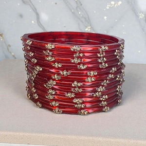 Op de afbeelding: Een stapel rode glazen armbanden met goudkleurige bloemversieringen. De armbanden zijn in een cirkelvormige opstelling gerangschikt, waarbij hun glanzende afwerking en ingewikkelde ontwerp worden getoond. De sieraden staan tegen een neutrale achtergrond.