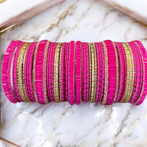 Op de afbeelding: Een stapel felroze armbanden met gouden accenten. De armbanden zijn in een cilindrische vorm gerangschikt en tonen een mix van effen roze en goudkleurige, mogelijk kralen, ontwerpen. De sieraden liggen op een marmerachtig oppervlak.
