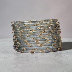 Op de afbeelding: Een stapel van tien doorzichtige, cilindrische armbanden met ingebedde goudkleurige glitterdeeltjes. Elke armband heeft een glad, afgerond oppervlak en een uniforme dikte, wat een samenhangende set van cirkelvormige sieraden vormt.