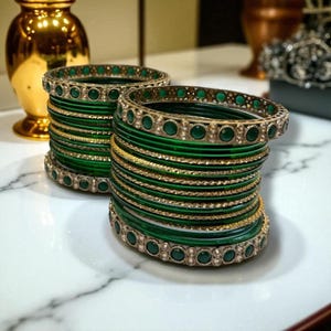 May include: Two stacks of green bangles with gold accents and decorative green stones. The bangles are stacked on a white marble surface, with a gold vase in the background. The bangles are a traditional jewelry item.