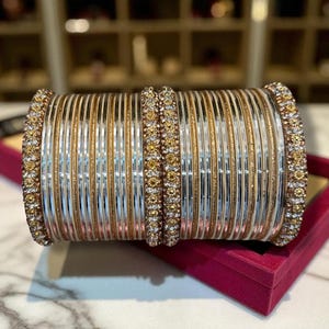 May include: A collection of gold and silver bangles with rhinestone detailing. The bangles are stacked, with alternating gold and silver bands. The edges of the bangles are adorned with rhinestones. The bangles are presented in a red box.