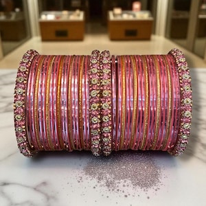 May include: A stack of pink and gold bangles with rhinestone accents. The bangles are arranged in a neat stack, with a scattering of glitter in the foreground. The bangles are a mix of solid pink and gold, with rhinestone embellishments.