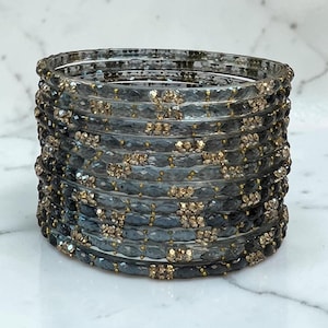 May include: A stack of multiple translucent dark grey and black bangles featuring a faceted texture. Each bangle is accented with small, shimmering gold-toned beads embedded at regular intervals along the circular band, creating a repeating pattern.