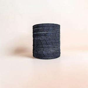 Puede incluir: Una pila de pulseras azul oscuro. Las pulseras están hechas de un material texturizado y están apiladas en forma cilíndrica. Las pulseras tienen un patrón sutil y se muestran sobre un fondo liso.