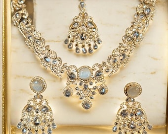 Könnte beinhalten: Ein goldfarbenes Schmuckset mit Halskette, Ohrringen und Kopfschmuck. Das Set ist mit klaren und grauen Edelsteinen verziert, die in aufwendigen Blumen- und Tropfenmustern angeordnet sind. Der Schmuck wird in einer goldgerahmten Box präsentiert.