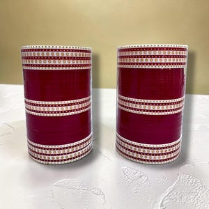 May include: Two cylindrical, maroon objects with white and gold decorative bands. The bands feature a repeating pattern of small gold floral designs. The objects are set against a neutral background.